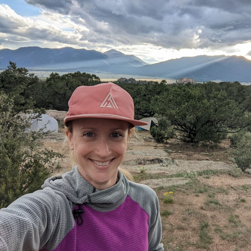 A woman with blonde hair smiles at the camera, wearing a pink hat and a purple and grey top. She appears to be outdoors, possibly camping, with tents visible in the background. The landscape includes trees and mountains under a cloudy sky, with sunlight breaking through.